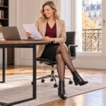 Une femme adulte blonde en blazer beige et jupe noire est assise à son bureau, jambes croisées, avec des bas noirs et des bottines.