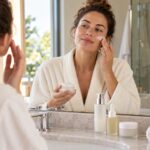 Femme adulte appliquant une crème sur sa joue devant un miroir, avec trois flacons de soin sur le plan de toilette.
